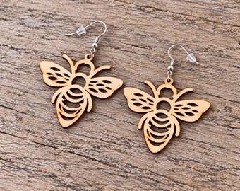 May include: A pair of dangle earrings featuring laser-cut wooden bee charms with intricate openwork details on the wings and abdomen. The light brown wooden bee shapes are suspended from silver-toned metal fishhook wires with clear plastic backings.