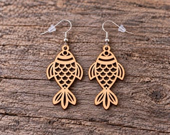 May include: A pair of light brown wooden fish earrings featuring a laser-cut scale pattern and a detailed tail design. Each fish pendant hangs from a silver-toned metal hook with a small clear plastic backing for secure placement on the ear.