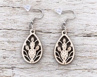 May include: A pair of teardrop-shaped earrings featuring a laser-cut wooden design of a prickly pear cactus. The earrings have silver-toned metal hooks with clear plastic backings, showcasing a light wood finish with dark brown engraved details.