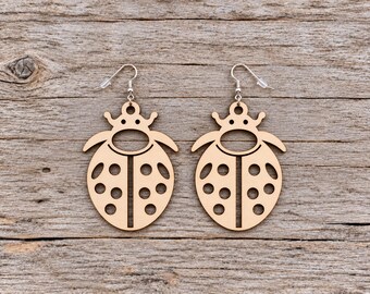 May include: A pair of light-colored wooden earrings shaped like ladybugs. Each earring features a cutout design with a central vertical line, circular holes representing spots, and a rounded top section with a small loop for the silver-toned metal hook.