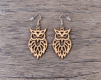 May include: A pair of dangle earrings featuring laser-cut wooden owl pendants with intricate geometric feather patterns. The light brown wooden owl shapes are suspended from silver-toned metal fishhook wires with clear plastic earring backs.