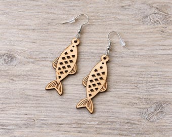 May include: A pair of dangle earrings featuring light brown wooden fish-shaped pendants. Each fish has a laser-cut diamond pattern on the body, a small circular eye, and etched fin and tail details. The pendants hang from silver-toned metal hooks.
