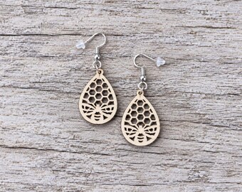 May include: A pair of drop earrings featuring light-colored wooden pendants in a teardrop shape. Each pendant displays a laser-cut design of a honeybee at the base with a honeycomb pattern above, suspended from silver-toned metal fishhook wires.