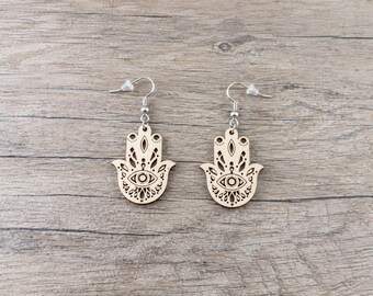 May include: A pair of light-colored wooden earrings shaped like Hamsa hands. Each earring features a laser-cut design of an eye in the center, surrounded by intricate floral and geometric patterns, suspended from silver-toned metal earring hooks.