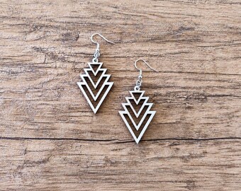 May include: A pair of dangle earrings featuring white, geometric, layered triangle cutouts. Each earring is suspended from a silver-toned metal hook. The design consists of four nested, open-frame triangles that decrease in size toward the top.