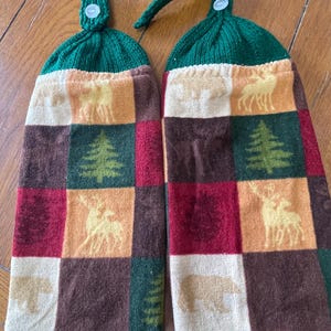 May include: Two hanging kitchen towels with a green knitted top and button closure. The towels feature a patchwork design with squares of brown, red, orange, and cream, each with a forest animal or tree silhouette.