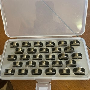May include: A clear plastic storage case filled with 36 silver bobbins for a sewing machine.
