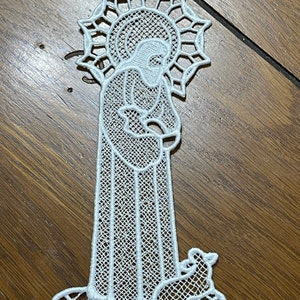 May include: White lace embroidery of a religious figure with a halo and a small bird at their feet.