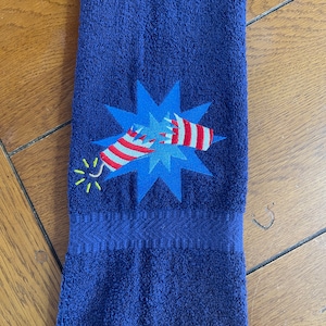 May include: A navy blue hand towel with an embroidered design of a red, white, and blue firework exploding in a blue starburst.