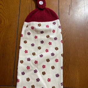 May include: A white kitchen towel with a red knitted hanging loop and a button. The towel has a pattern of colorful peppermint candies.