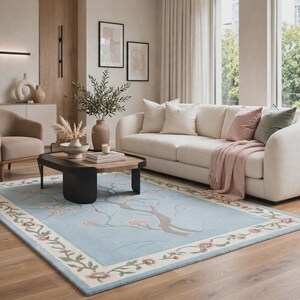 May include: A rectangular area rug featuring a light blue center with a brown tree branch design and pink blossoms. The rug has a wide cream-colored border decorated with a repeating pattern of pink flowers and green vines along the edges.