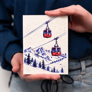 May include: A rectangular greeting card featuring a detailed illustration of two bright red cable cars suspended on dark blue cables above a snowy mountain landscape with dark blue pine trees and scattered red speckles on a cream background.