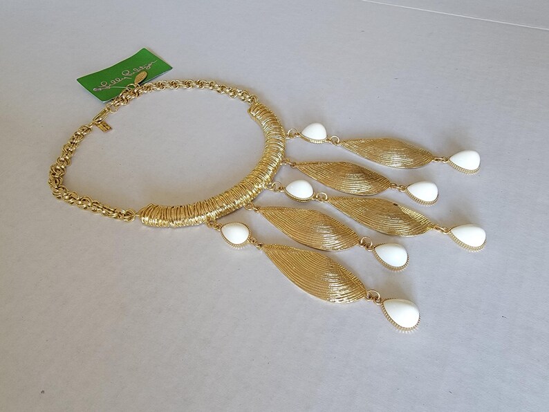 May include: Gold-tone statement necklace with a textured collar and chain. The necklace features five gold leaf-shaped pendants with white teardrop-shaped accents. A green tag is visible.