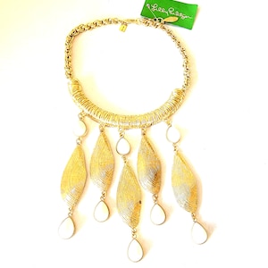 May include: Gold-toned statement necklace with a woven design and teardrop-shaped white accents. The necklace features a thick chain and a tag that reads "Lilly Pulitzer".