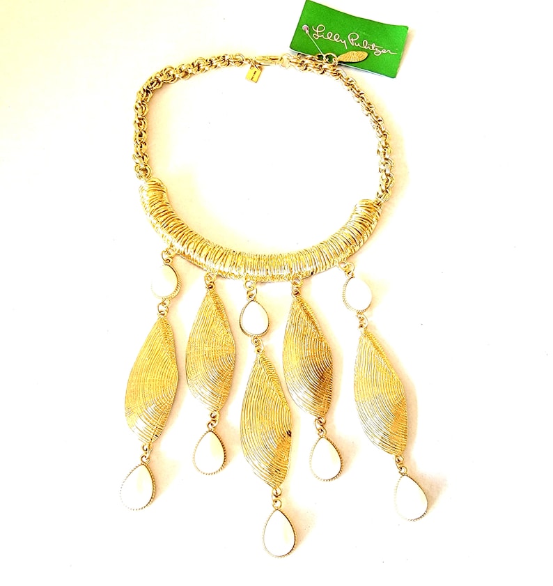 May include: Gold-toned statement necklace with a woven design and teardrop-shaped white accents. The necklace features a thick chain and a tag that reads "Lilly Pulitzer".