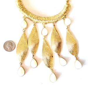 May include: Gold-toned statement necklace with a textured collar and dangling leaf-shaped pendants. Each pendant features a cream-colored, teardrop-shaped accent. The necklace is displayed on a white background, showcasing its intricate design.