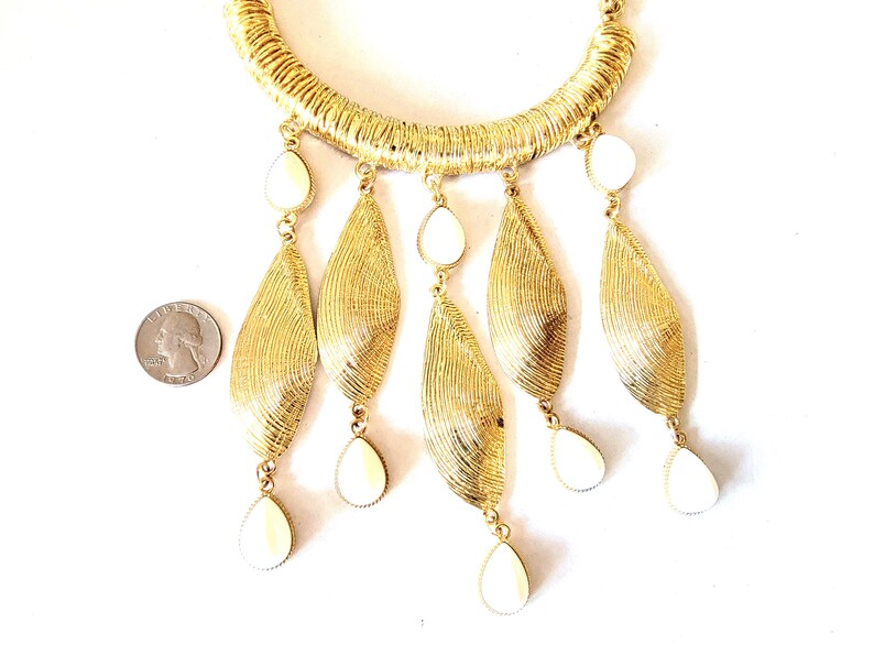 May include: Gold-toned statement necklace with a textured collar and dangling leaf-shaped pendants. Each pendant features a cream-colored, teardrop-shaped accent. The necklace is displayed on a white background, showcasing its intricate design.