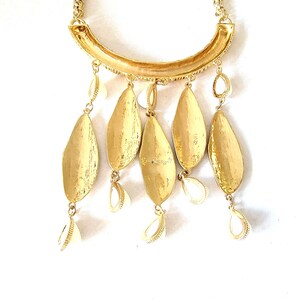 May include: Gold-toned statement necklace featuring a curved bar pendant with textured detailing. The necklace has five large, leaf-shaped pendants and smaller shell-shaped charms. The design is completed with a delicate chain.