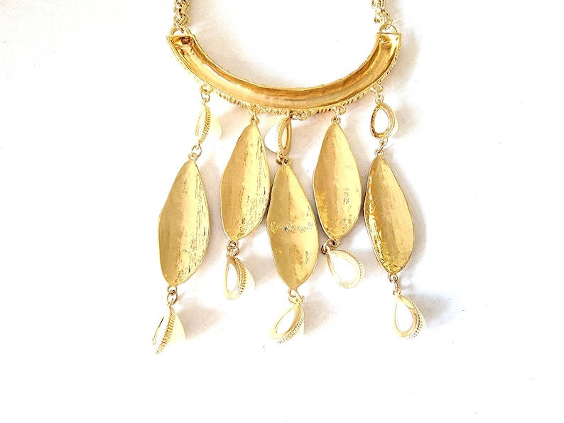 May include: Gold-toned statement necklace featuring a curved bar pendant with textured detailing. The necklace has five large, leaf-shaped pendants and smaller shell-shaped charms. The design is completed with a delicate chain.