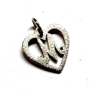 May include: Silver-toned pendant in the shape of a heart with the letter "M" inside. The pendant has a textured surface and a small loop at the top for a chain. The heart shape is open, allowing light to pass through.