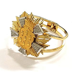 Puede incluir: Un brazalete dorado con un emblema central que presenta un diseño de escudo y corona. El emblema está rodeado de elementos dorados radiantes y acentuado con triángulos plateados con gemas. El brazalete tiene un cierre de bisagra.