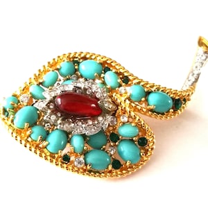 May include: A gold-toned brooch with a leaf-like design, adorned with turquoise cabochons, clear rhinestones, and a central red stone. The brooch has a textured gold border and a rhinestone-studded stem.