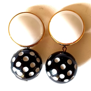 May include: A pair of statement earrings. Each earring features a large white disc with a gold-tone border, connected to a black sphere adorned with white polka dots. The earrings have a post-back closure.