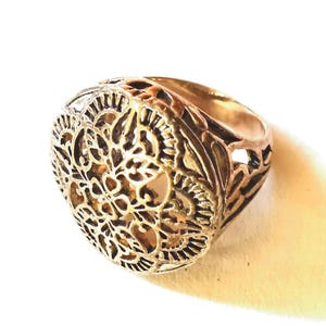 May include: A gold-toned metal ring featuring a large, circular face with an intricate, openwork filigree pattern. The design consists of symmetrical floral and leaf-like motifs that extend along the sides of the band, creating a detailed texture.