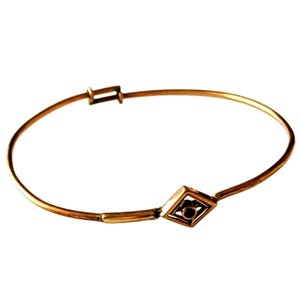 May include: A gold-colored bangle bracelet with a geometric diamond-shaped accent. The bracelet has a clasp closure and a simple, elegant design. The diamond-shaped accent features a dark center.