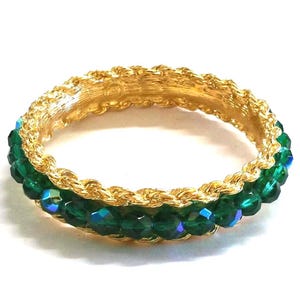 Puede incluir: Un anillo dorado con un diseño trenzado y una fila de cuentas verdes facetadas. El anillo tiene una superficie brillante y reflectante y está sobre un fondo blanco. Las cuentas tienen una ligera iridiscencia azul.