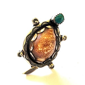 May include: A handcrafted silver turtle pendant featuring a natural shell center with brown and cream patterns. The pendant has a turquoise stone accent and intricate metalwork around the shell, with decorative elements on the legs and head.