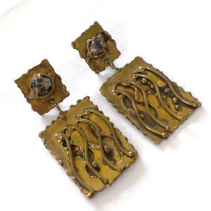 May include: A pair of brass-toned drop earrings featuring a square top component with a central dark stone and a larger rectangular bottom component decorated with raised, wavy, linear metal patterns and scalloped edges on all sides.