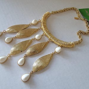 May include: Gold-tone statement necklace featuring a textured, ribbed collar and a chain. The necklace has five gold leaf-shaped pendants with white teardrop-shaped accents. A green tag with white text is attached.