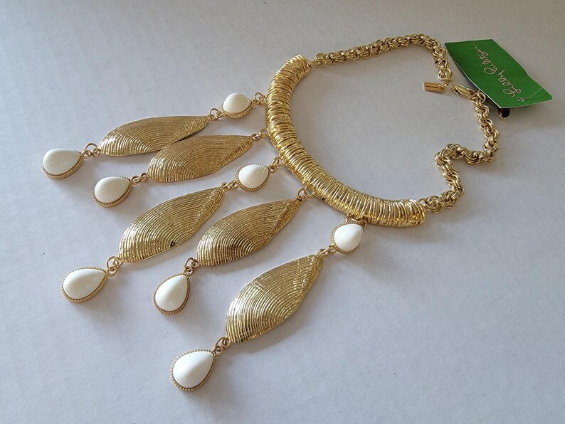 May include: Gold-tone statement necklace featuring a textured, ribbed collar and a chain. The necklace has five gold leaf-shaped pendants with white teardrop-shaped accents. A green tag with white text is attached.