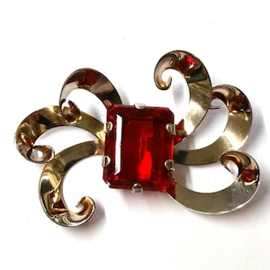 Könnte beinhalten: Vintage Brosche mit einem großen, rechteckigen, rubinroten Edelstein in der Mitte. Die Brosche hat ein silberfarbenes Metalldesign mit wirbelnden, bandartigen Formen. Der Edelstein ist in einen silbernen Rahmen gefasst.