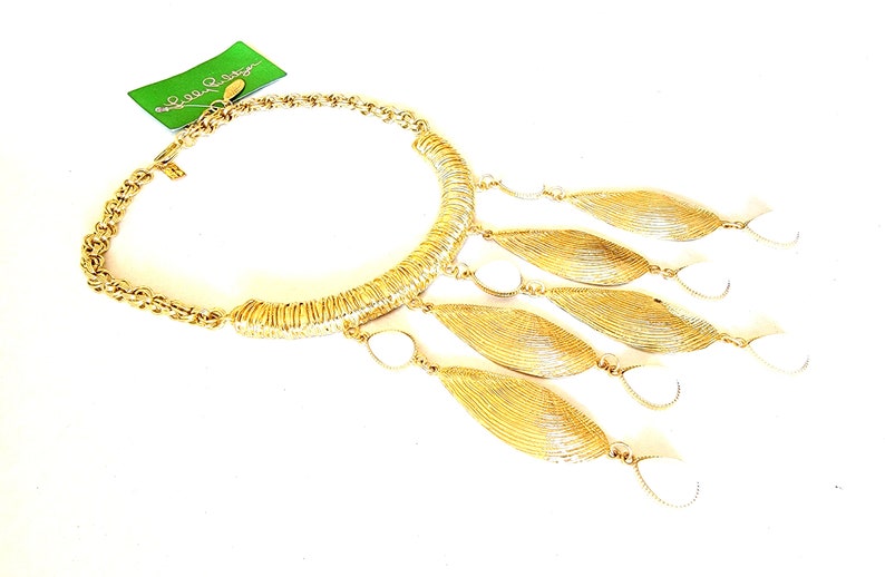 May include: Gold-tone necklace with a chain and a curved, textured centerpiece. The necklace features dangling leaf-shaped pendants with white accents. A green tag with the brand name is attached.