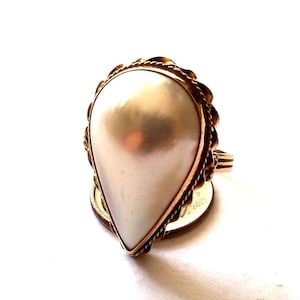 May include: A gold ring featuring a large, pear-shaped white pearl set within a decorative, twisted rope-style metal border. The ring is positioned directly on top of a circular gold-colored coin, highlighting the scale of the jewelry piece.