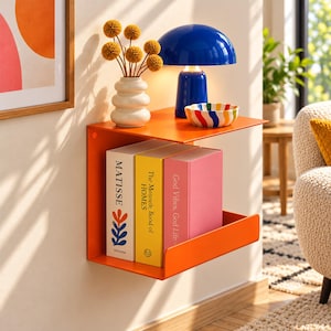 May include: A bright orange wall-mounted metal shelf holding three books titled Matisse, The Monocle Book of Homes, and God Vibes, God Life. On top sits a white wavy vase with yellow flowers, a blue mushroom-shaped lamp, and a small bowl.