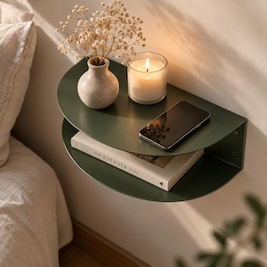 May include: A semi-circular dark green metal wall shelf with two tiers. The top shelf holds a small beige vase with dried flowers, a lit white candle in a glass jar, and a black smartphone. The bottom shelf holds a book titled The Kinfolk Home.