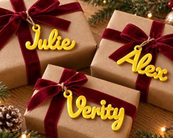 May include: Three brown paper-wrapped gift boxes, each tied with a deep red velvet ribbon. Attached to each ribbon is a bright yellow, custom-cut name tag featuring the names Julie, Alex, and Verity in a cursive, rounded script font.