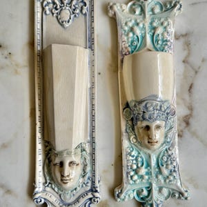 May include: Two vintage ceramic wall pockets with ornate blue and white designs. Each pocket features a sculpted face and a decorative floral pattern.
