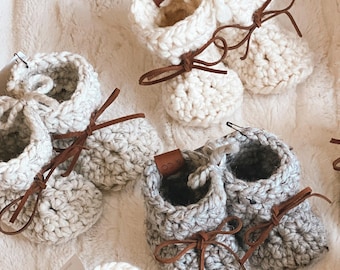 May include: A set of crocheted baby booties in various colours, including white, grey, black, and yellow. The booties have brown leather laces and are arranged on a white fluffy surface. A crocheted sunflower is also included in the image.