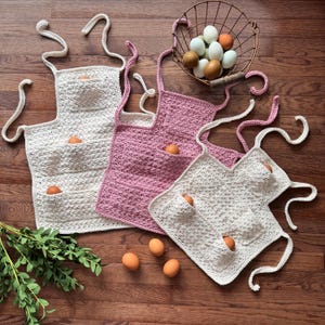 May include: Three handmade crochet egg gathering aprons in cream and pink, each with multiple pockets. Brown eggs are nestled in the pockets and scattered on the wooden surface. A wire basket filled with eggs sits in the background.