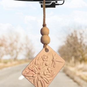 May include: A rectangular, light-coloured, textured clay ornament hanging from a rearview mirror by a brown twine string with two round wooden beads. The ornament features a raised relief design of a cluster of flowers and a dragonfly.
