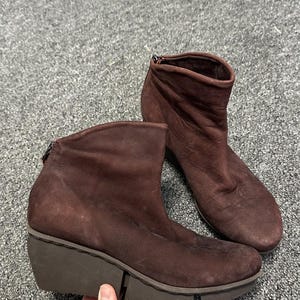May include: A pair of dark brown ankle boots featuring a soft, matte finish, a rounded toe, and a thick, dark brown rubber wedge sole. The boots have a zip closure at the back of the heel and a simple, unadorned design with visible stitching.
