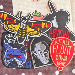May include: A collection of five embroidered patches featuring a moth with a skull pattern, a silhouette with the text PSYCHO, a cat with red eyes and the text PET SEMATARY, a portrait in an arch, and a red balloon with WE ALL FLOAT DOWN HERE.