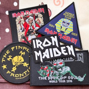 Puede incluir: Una colección de cuatro parches bordados de Iron Maiden. Un parche cuadrado muestra una figura en rojo, uno triangular exhibe una máscara de faraón con texto, uno circular presenta una calavera y uno rectangular muestra una criatura verde.