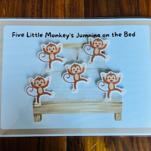 Pode incluir: Um cartão educativo plastificado com o texto Five Little Monkey's Jumping on the Bed. O cartão apresenta um gráfico de uma estrutura de cama de madeira com roupa de cama branca e cinco macacos castanhos em poses de salto.