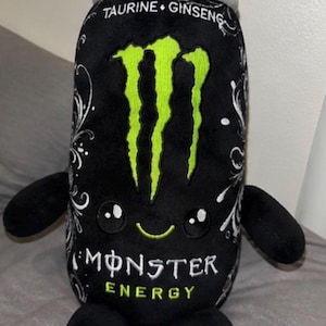 May include: A black plush toy shaped like an energy drink can with a bright green claw logo, white floral accents, and the text TAURINE GINSENG and MONSTER ENERGY. It features a smiling face with two black eyes, two arms, and two small feet.
