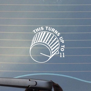 May include: White vinyl decal featuring a stylized volume knob with the text "THIS TURNS UP TO 11". The design is a simple, bold graphic, suitable for application on a car window or other smooth surfaces.